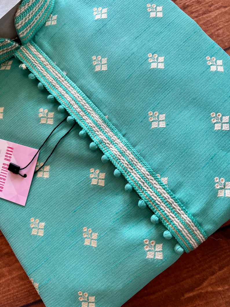Pode incluir: Uma kurta azul turquesa com um padr&atilde;o bordado branco e dourado. A kurta tem uma bainha branca com detalhes dourados e pompons brancos ao longo das bordas. A etiqueta diz "KASH COLLECTION".