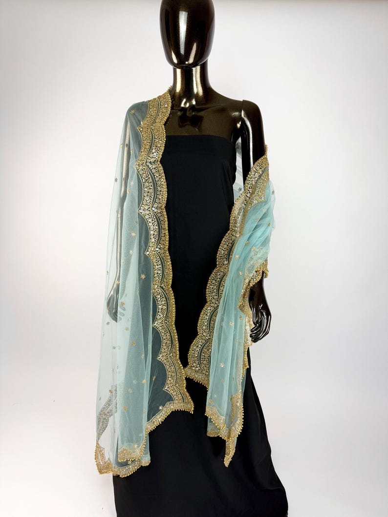 May include: A pale blue sheer dupatta with gold embroidery and embellishments. The dupatta is draped over a black dress on a mannequin. The gold trim has a scalloped edge. The dupatta has small gold dots throughout.