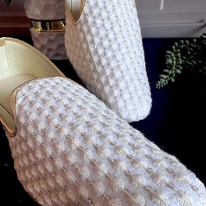 Pode incluir: Um par de chinelos brancos bordados com acabamento dourado. Os chinelos t&ecirc;m bico quadrado e sola plana.