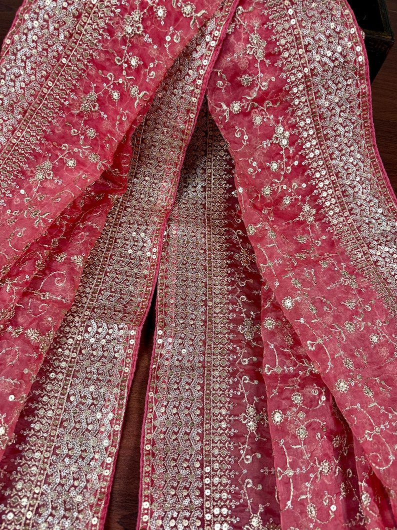 Peut inclure: Un tissu rose vif et transparent, orn&eacute; de broderies florales et de sequins argent&eacute;s. Le tissu est drap&eacute;, mettant en valeur les d&eacute;tails des ornements et la texture d&eacute;licate. Le design pr&eacute;sente un panneau central et des bordures.