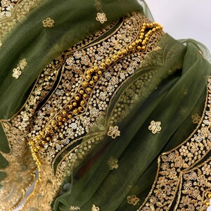 May include: Close-up of a green fabric with intricate gold embroidery and embellishments. The design features floral patterns, sequins, and beaded accents. The fabric is layered, creating depth and texture, with a sheer overlay.