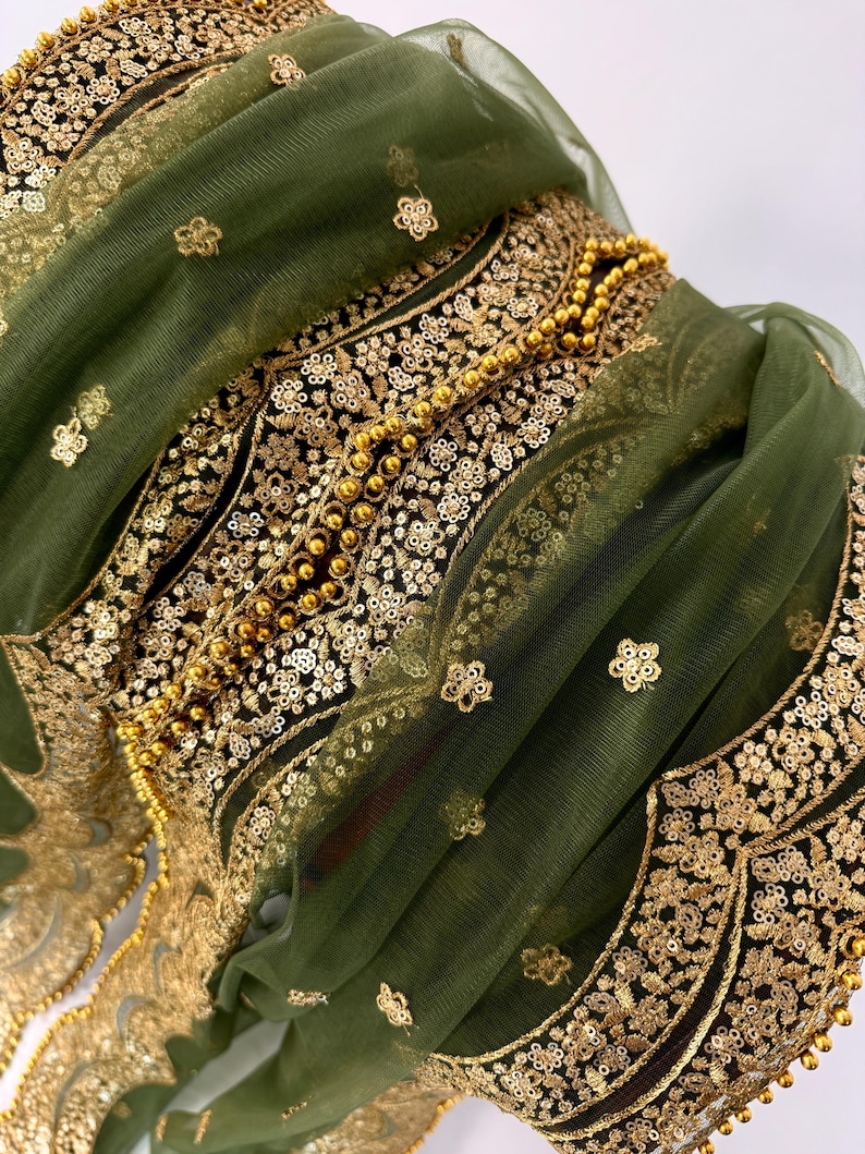 May include: Close-up of a green fabric with intricate gold embroidery and embellishments. The design features floral patterns, sequins, and beaded accents. The fabric is layered, creating depth and texture, with a sheer overlay.