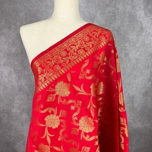 May include: A vibrant red fabric with an intricate gold floral pattern. The fabric is draped over a mannequin, showcasing the detailed design. The border features a repeating gold floral motif. This textile is suitable for various sewing projects.