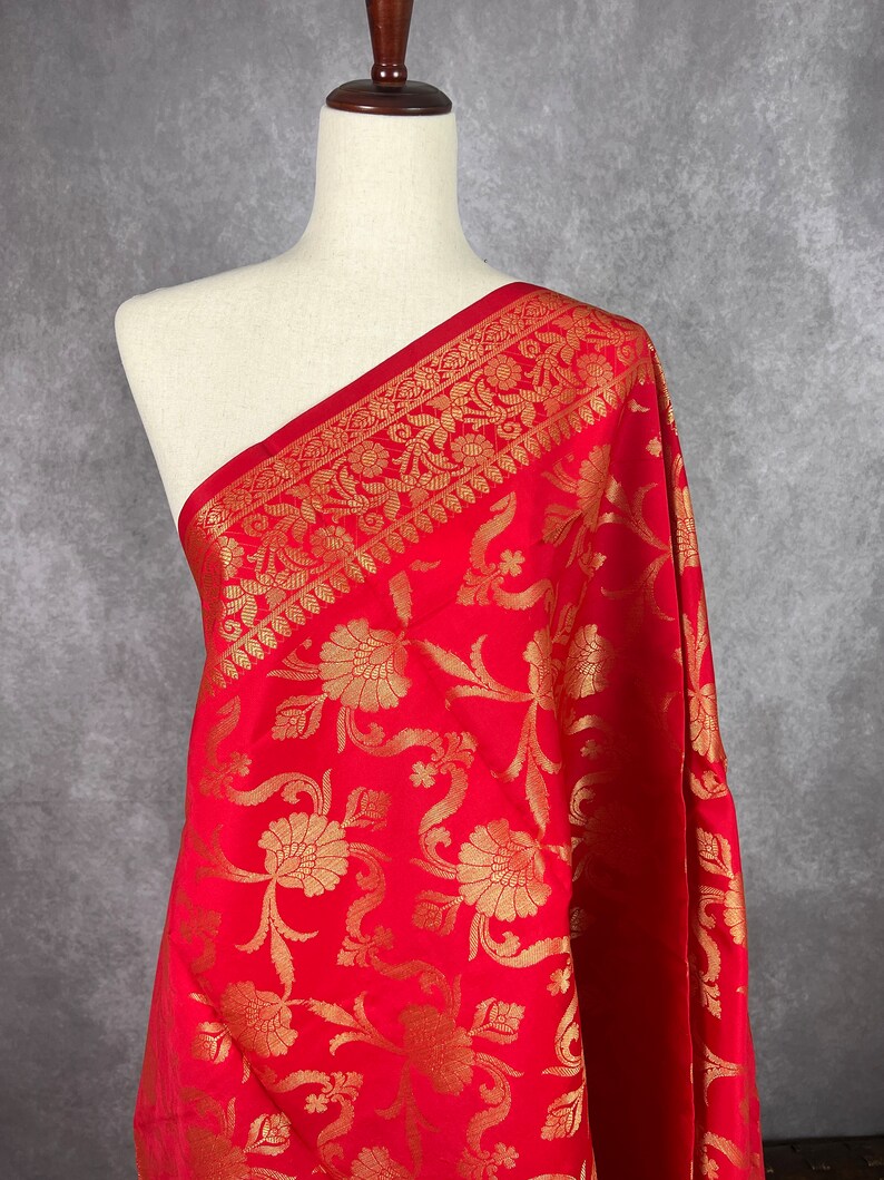 May include: A vibrant red fabric with an intricate gold floral pattern. The fabric is draped over a mannequin, showcasing the detailed design. The border features a repeating gold floral motif. This textile is suitable for various sewing projects.