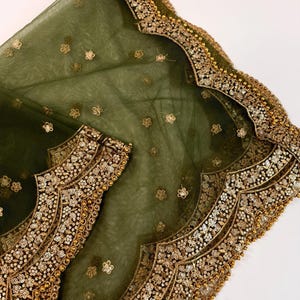 May include: Olive green sheer fabric with gold floral embroidery and scalloped edges. The fabric is adorned with small, scattered gold flower-shaped sequins and larger, ornate gold embellishments along the borders. The design suggests a formal garment or accessory.