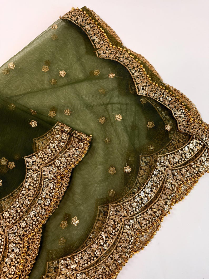 May include: Olive green sheer fabric with gold floral embroidery and scalloped edges. The fabric is adorned with small, scattered gold flower-shaped sequins and larger, ornate gold embellishments along the borders. The design suggests a formal garment or accessory.