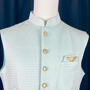 May include: A light blue sleeveless vest with a white geometric pattern and gold buttons. The vest has a pocket on the right side with a gold embroidered design.