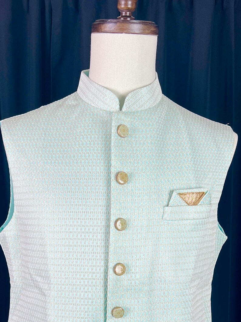May include: A light blue sleeveless vest with a white geometric pattern and gold buttons. The vest has a pocket on the right side with a gold embroidered design.