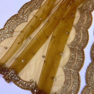 May include: A sheer, golden-yellow fabric with a scalloped edge, embellished with sequins and small, sparkling accents. The fabric is draped over a cream-colored base, creating a layered effect. The border is intricately detailed with gold beads and sequins.