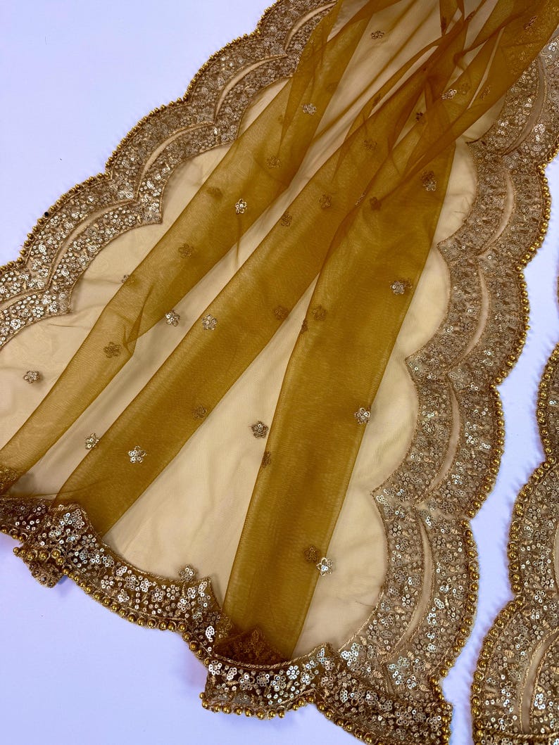 May include: A sheer, golden-yellow fabric with a scalloped edge, embellished with sequins and small, sparkling accents. The fabric is draped over a cream-colored base, creating a layered effect. The border is intricately detailed with gold beads and sequins.