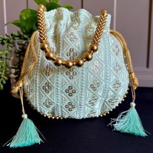 May include: A light blue drawstring pouch with gold embroidery and a beaded handle. The pouch features a diamond pattern and is accented with gold trim and turquoise tassels. The bag is approximately 20 cm in height.