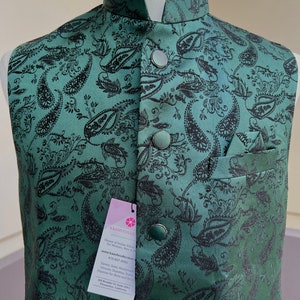 May include: A green paisley patterned waistcoat with black buttons and a pocket on the right side. The waistcoat has a mandarin collar and is made of a silky fabric. The tag on the waistcoat reads "Kaash Collection, House of Indian Ethnic Wear for Women, Men & Kids, www.kaashcollection.com, 415-827-9432, Sarees, Suits, Readymade Blouses, Dupattas, Kurta Pajamas for Mens and Dresses, 405 Boddie Ct Suite 209, Pleasanton, California."