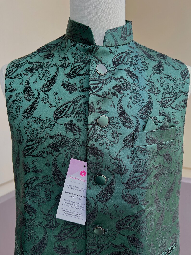 May include: A green paisley patterned waistcoat with black buttons and a pocket on the right side. The waistcoat has a mandarin collar and is made of a silky fabric. The tag on the waistcoat reads "Kaash Collection, House of Indian Ethnic Wear for Women, Men & Kids, www.kaashcollection.com, 415-827-9432, Sarees, Suits, Readymade Blouses, Dupattas, Kurta Pajamas for Mens and Dresses, 405 Boddie Ct Suite 209, Pleasanton, California."