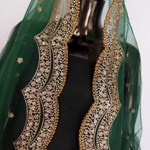 Peut inclure: Un ch&acirc;le vert fonc&eacute; transparent avec des broderies dor&eacute;es et des d&eacute;tails de paillettes. Le ch&acirc;le pr&eacute;sente une bordure d&eacute;corative avec des motifs floraux et de petites perles dor&eacute;es. Le v&ecirc;tement est drap&eacute; sur une forme noire, mettant en valeur le design.