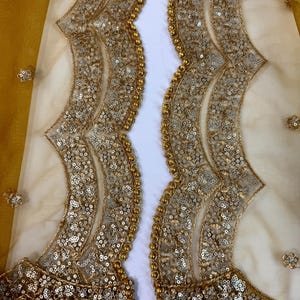 May include: Close-up of a sheer, cream-colored fabric with intricate gold sequin and bead embellishments. The design features a scalloped edge and floral accents, creating a luxurious and ornate look. The fabric has a yellow-gold border.