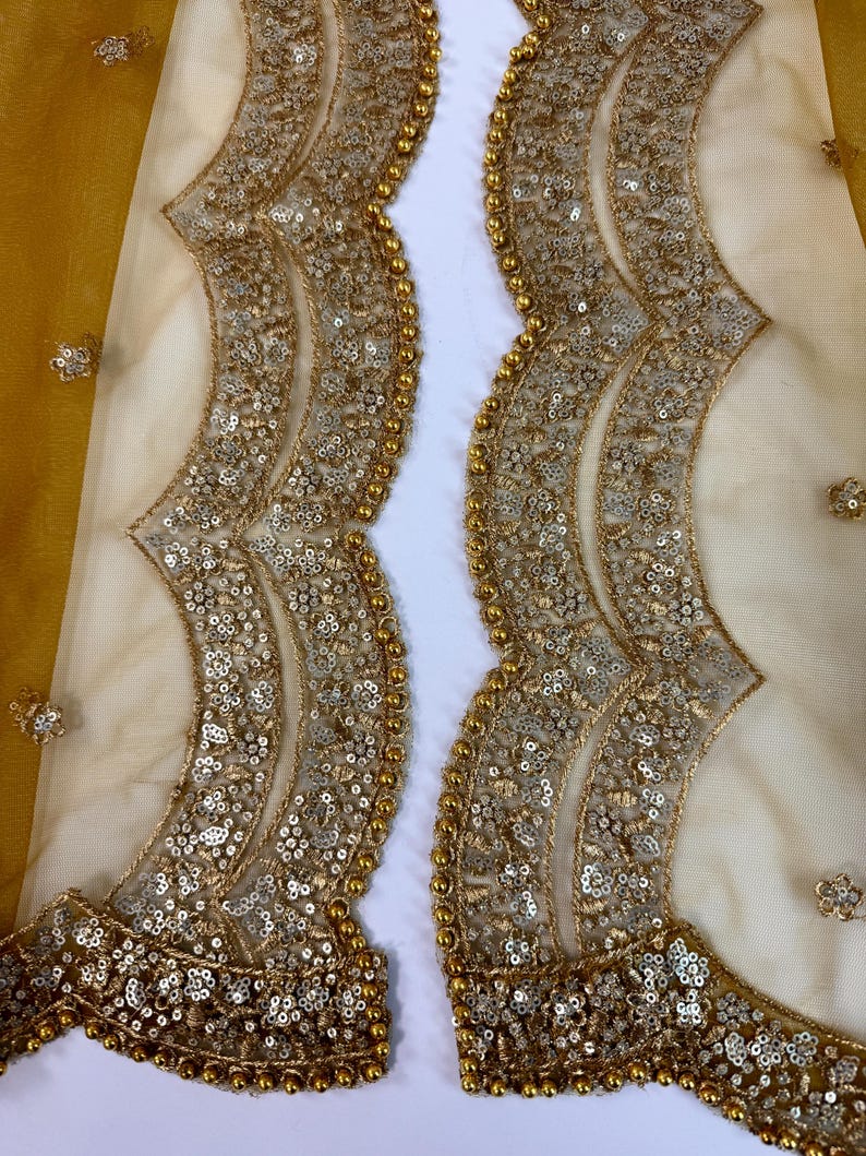 May include: Close-up of a sheer, cream-colored fabric with intricate gold sequin and bead embellishments. The design features a scalloped edge and floral accents, creating a luxurious and ornate look. The fabric has a yellow-gold border.