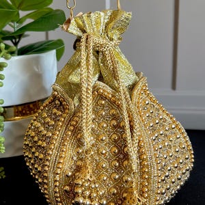 May include: A gold-colored, beaded drawstring bag with a decorative tassel. The bag is embellished with small, round beads and intricate patterns. The bag has a gold-colored handle and is likely used as a clutch or small purse.
