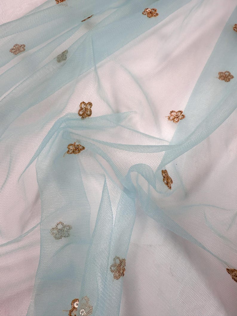 May include: Pale blue tulle fabric with gold floral embroidery. The sheer material is draped, creating soft folds. The delicate embroidery adds a touch of elegance to the fabric, suitable for craft projects or clothing. The fabric is lightweight and airy.