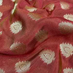 May include: Close-up of a draped fabric with a reddish-brown hue. The fabric features a repeating pattern of gold-colored leaf-shaped designs. The material appears soft and has a slight sheen, suggesting a luxurious textile.