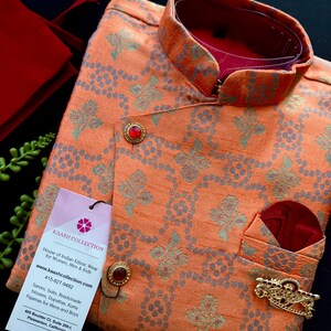 May include: An orange and gray patterned kurta set with a mandarin collar and red accents. The set includes a matching pocket square and decorative gold brooch. A tag from Kaash Collection is attached.