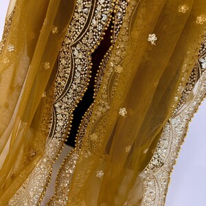 May include: Mustard yellow sheer fabric with intricate gold embroidery and embellishments. The design features floral patterns and a beaded border. The fabric is draped, showcasing the delicate texture and detailed craftsmanship. This textile is suitable for special occasion wear.