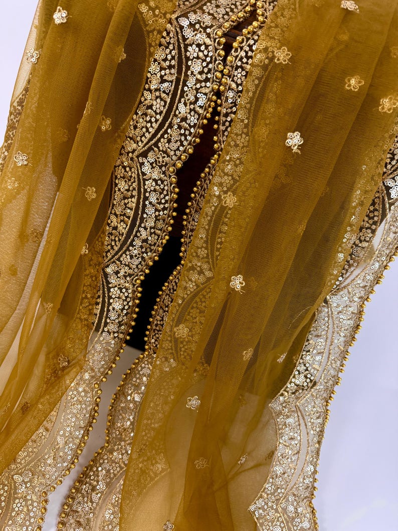 May include: Mustard yellow sheer fabric with intricate gold embroidery and embellishments. The design features floral patterns and a beaded border. The fabric is draped, showcasing the delicate texture and detailed craftsmanship. This textile is suitable for special occasion wear.