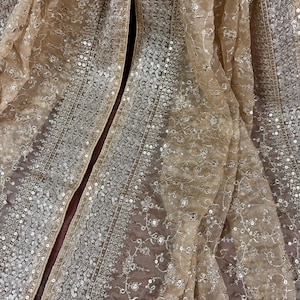 May include: Close-up of a beige, sheer fabric with intricate embroidery and sequin embellishments. The fabric is draped, revealing detailed patterns and textures. The design features floral and geometric motifs, creating a luxurious and elegant appearance.