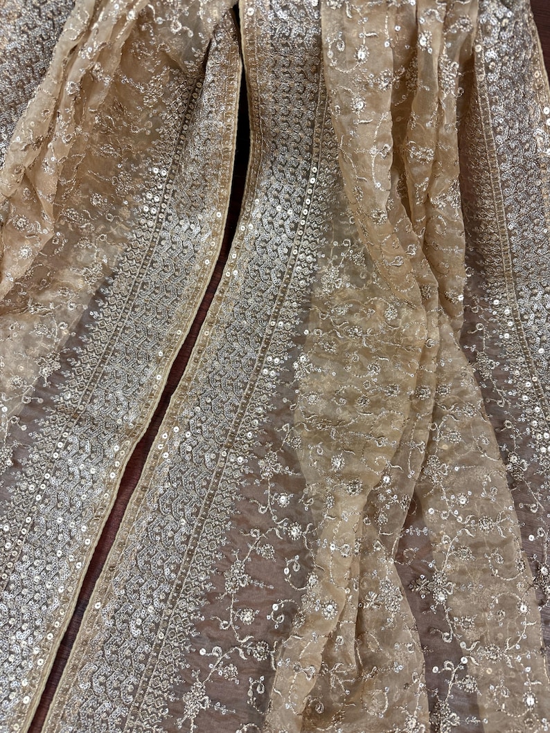 May include: Close-up of a beige, sheer fabric with intricate embroidery and sequin embellishments. The fabric is draped, revealing detailed patterns and textures. The design features floral and geometric motifs, creating a luxurious and elegant appearance.