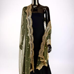 May include: Olive green sheer shawl with gold embroidered trim and small gold embellishments. The shawl is draped over a black dress on a mannequin. The gold trim features a detailed, ornate design.