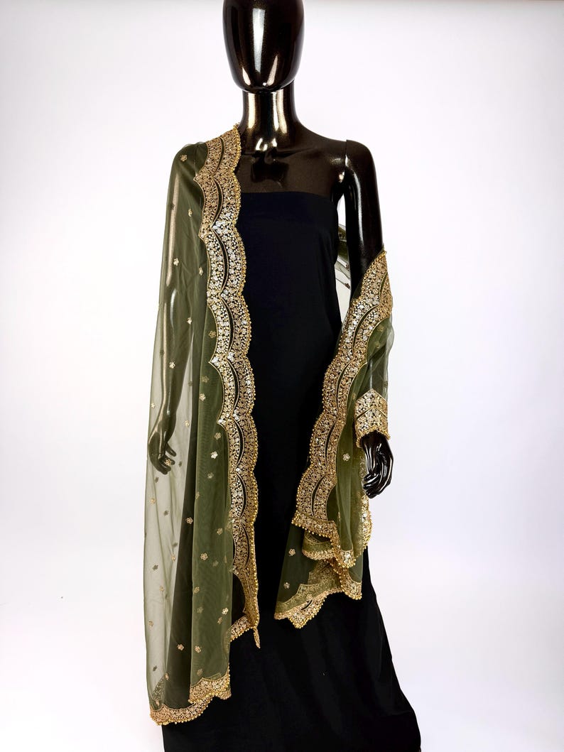 May include: Olive green sheer shawl with gold embroidered trim and small gold embellishments. The shawl is draped over a black dress on a mannequin. The gold trim features a detailed, ornate design.
