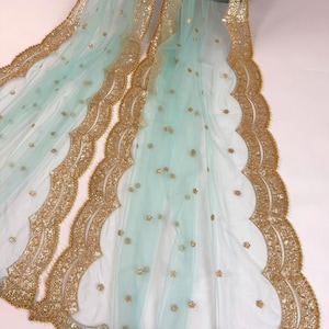 May include: A delicate, pale blue fabric with gold detailing. The edges are adorned with a scalloped gold border, and small gold floral accents are scattered across the surface. The fabric is draped, highlighting its flowing texture.