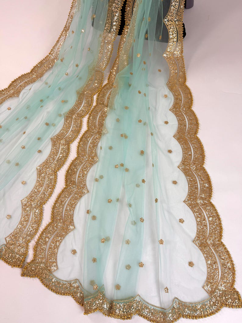May include: A delicate, pale blue fabric with gold detailing. The edges are adorned with a scalloped gold border, and small gold floral accents are scattered across the surface. The fabric is draped, highlighting its flowing texture.