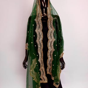 Peut inclure: Un ch&acirc;le vert fonc&eacute; transparent avec broderies et ornements dor&eacute;s. Le ch&acirc;le est drap&eacute; sur un mannequin noir, mettant en valeur le design et les d&eacute;tails complexes. Les bords sont orn&eacute;s d'or, ajoutant une touche d'&eacute;l&eacute;gance.