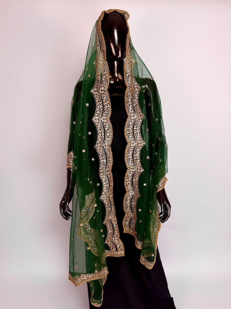 Peut inclure: Un ch&acirc;le vert fonc&eacute; transparent avec broderies et ornements dor&eacute;s. Le ch&acirc;le est drap&eacute; sur un mannequin noir, mettant en valeur le design et les d&eacute;tails complexes. Les bords sont orn&eacute;s d'or, ajoutant une touche d'&eacute;l&eacute;gance.