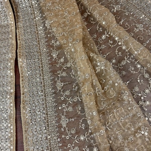 May include: Close-up of a sheer, beige fabric with intricate silver sequin and embroidered floral detailing. The fabric is draped, showcasing the delicate texture and design. The edges are trimmed with a silver sequin border.