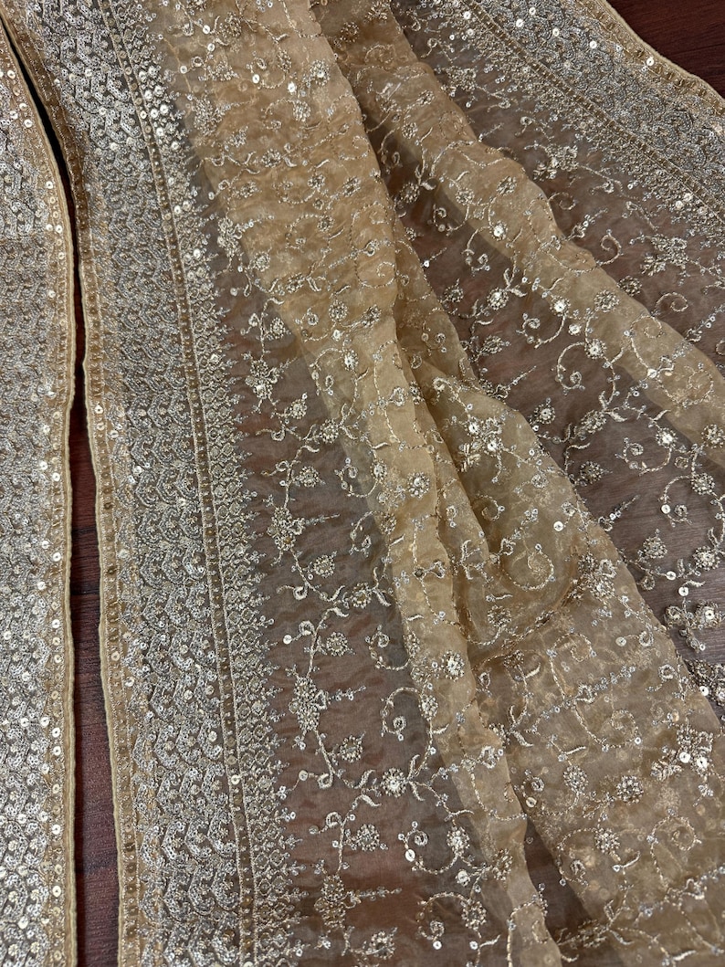 May include: Close-up of a sheer, beige fabric with intricate silver sequin and embroidered floral detailing. The fabric is draped, showcasing the delicate texture and design. The edges are trimmed with a silver sequin border.