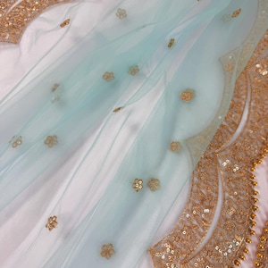 May include: A delicate, pale blue fabric with gold floral embroidery and a scalloped gold sequin border. The fabric is draped over a white surface, creating soft folds. Gold embellishments add a touch of elegance to the design.