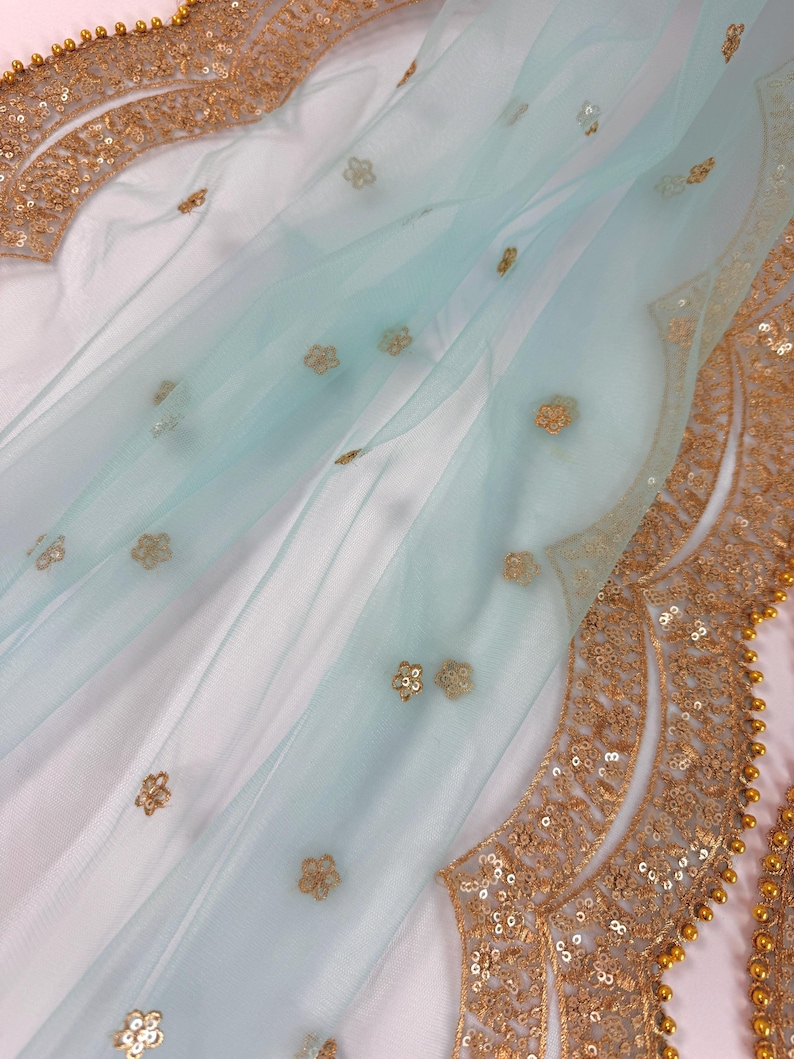 May include: A delicate, pale blue fabric with gold floral embroidery and a scalloped gold sequin border. The fabric is draped over a white surface, creating soft folds. Gold embellishments add a touch of elegance to the design.