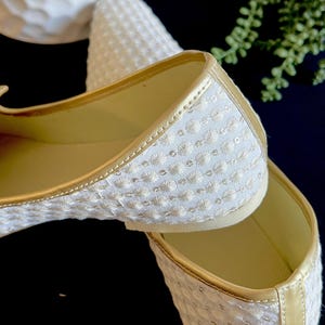 Pode incluir: Um par de sapatos baixos brancos bordados com detalhes dourados. Os sapatos s&atilde;o feitos de um tecido macio e texturizado e t&ecirc;m um ajuste confort&aacute;vel.