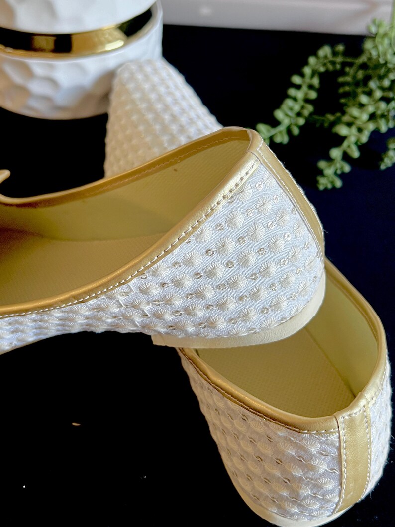 Pode incluir: Um par de sapatos baixos brancos bordados com detalhes dourados. Os sapatos s&atilde;o feitos de um tecido macio e texturizado e t&ecirc;m um ajuste confort&aacute;vel.