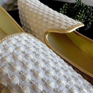 Pode incluir: Close-up de um par de chinelos brancos bordados com guarni&ccedil;&otilde;es douradas. Os chinelos t&ecirc;m um padr&atilde;o floral delicado.