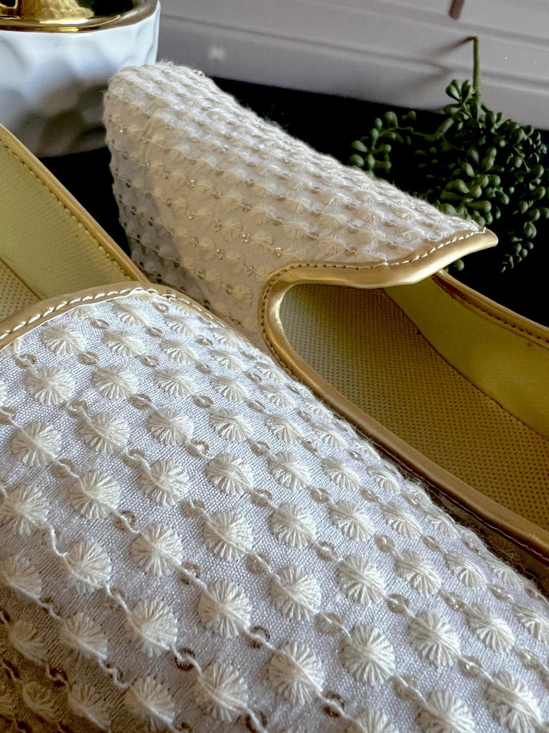Pode incluir: Close-up de um par de chinelos brancos bordados com guarni&ccedil;&otilde;es douradas. Os chinelos t&ecirc;m um padr&atilde;o floral delicado.