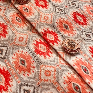 May include: Close-up of a white fabric with a red, gray, and white geometric pattern. The fabric has two gold-toned buttons with clear rhinestones.