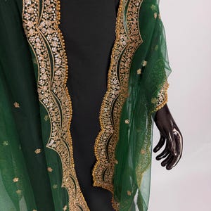 Peut inclure: Un ch&acirc;le vert &eacute;meraude transparent avec des broderies dor&eacute;es et des sequins d&eacute;taill&eacute;s sur les bords. Le ch&acirc;le est drap&eacute; sur un v&ecirc;tement noir, mettant en valeur le contraste. Les ornements dor&eacute;s ajoutent une touche d'&eacute;l&eacute;gance.