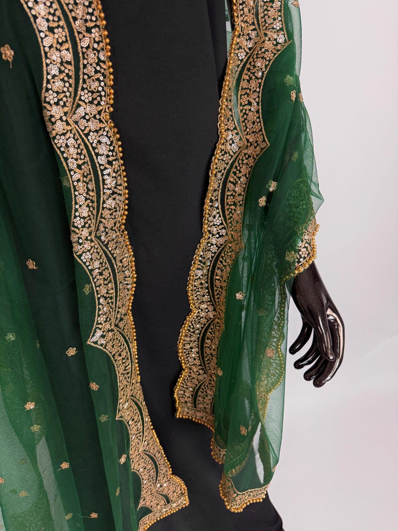 Peut inclure: Un ch&acirc;le vert &eacute;meraude transparent avec des broderies dor&eacute;es et des sequins d&eacute;taill&eacute;s sur les bords. Le ch&acirc;le est drap&eacute; sur un v&ecirc;tement noir, mettant en valeur le contraste. Les ornements dor&eacute;s ajoutent une touche d'&eacute;l&eacute;gance.