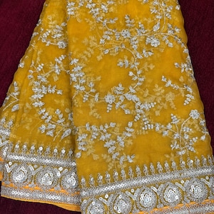 May include: A yellow sheer fabric saree with silver embroidery and sequins. The saree has a floral design and a border with a silver and orange trim.