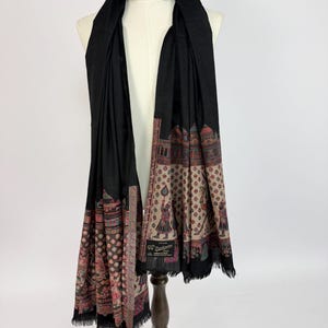 May include: A black scarf featuring a detailed border pattern with beige, red, and blue colors. The design includes small figures and architectural motifs. The scarf has a fringed edge and a small black label with the text Pashmina.