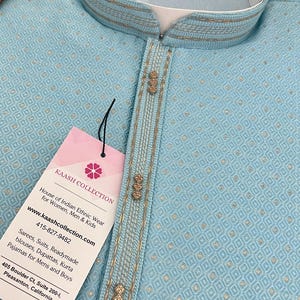 May include: A light blue kurta with gold trim and decorative buttons. The fabric has a subtle diamond pattern. The tag reads "KAASH COLLECTION House of Indian Ethnic Wear for Women, Men & Kids www.kaashcollection.com 415-827-9482 Sarees, Suits, Readymade blouses, Dupattas, Kurta Pajamas for Mens and Boys 405 Boulder Ct, Suite 200-1, Pleasanton, California"