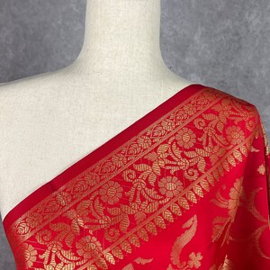 May include: A vibrant red fabric with an ornate gold floral border is draped over a mannequin. The gold pattern features detailed flowers and leaves. The fabric's rich color and intricate design suggest a luxurious textile.
