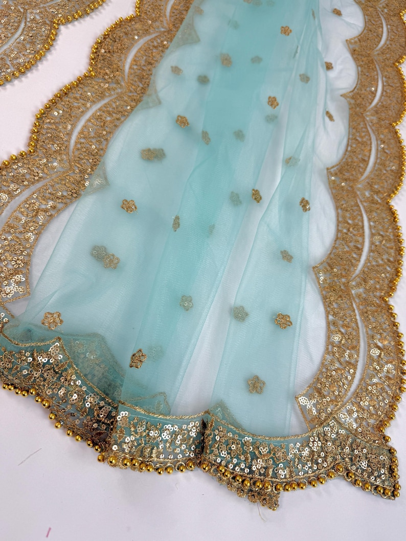 May include: A light blue sheer fabric with a scalloped gold sequin border. The fabric features small gold floral embellishments. The border is detailed with gold sequins and small gold beads.